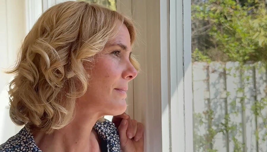 An older woman with glasses, captured in a half-profile, is sitting on a fabric sofa and smiling.