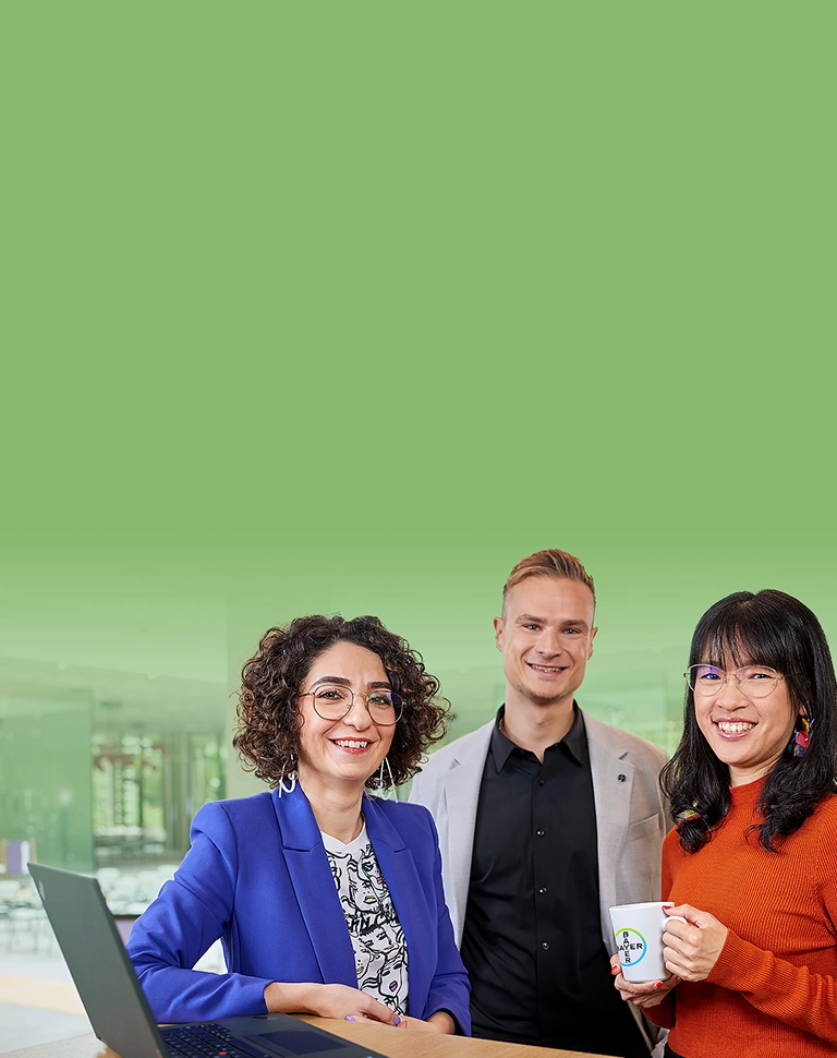 Two women and one man in business attire are posing and smiling at the camera. From left to right: a woman wearing a blue blazer and glasses, holding a laptop; a man in a black shirt and beige blazer; and a woman in a red blouse holding a cup with the Bayer logo. An office environment is visible in the background.