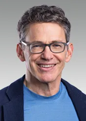 Bill Anderson is wearing a light blue shirt, a blue blazer, and glasses, and is smiling at the camera.