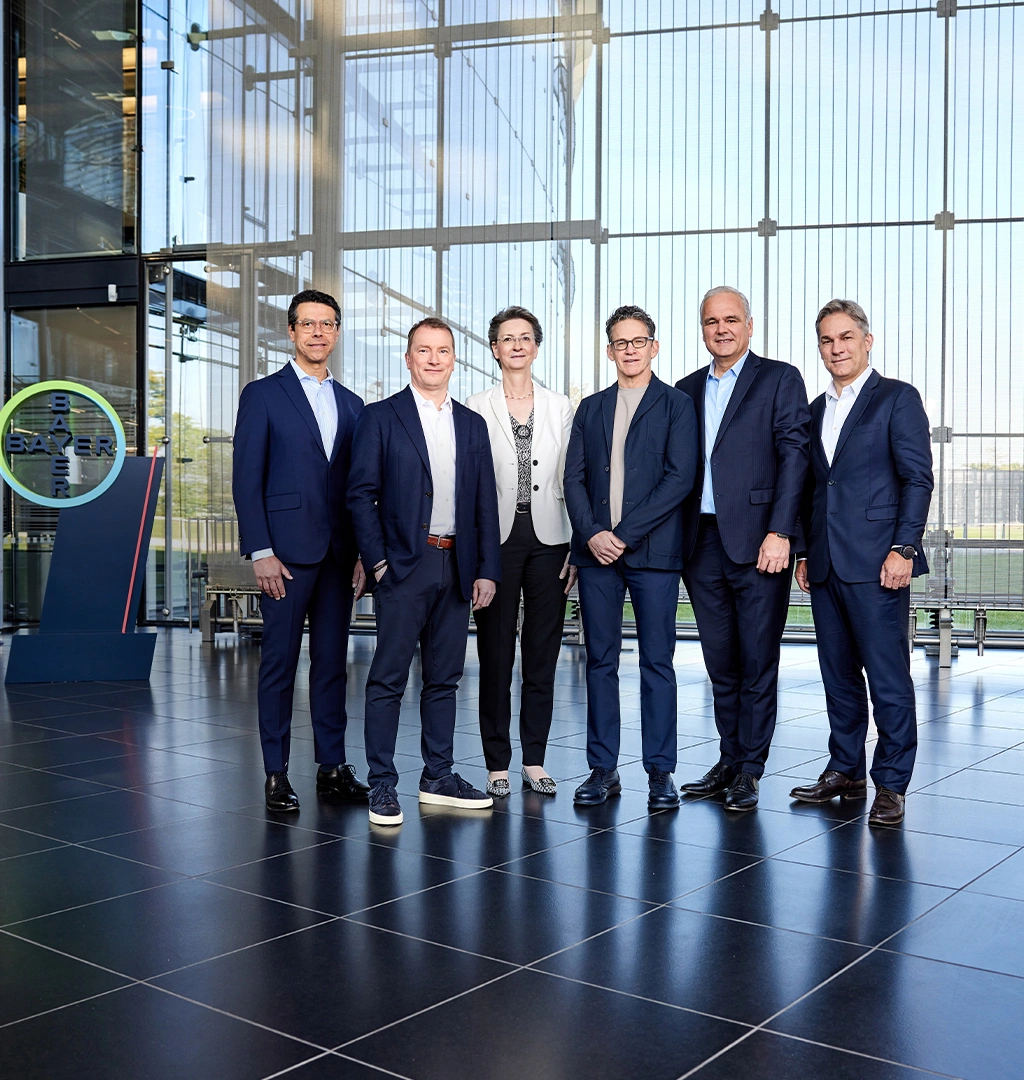 The board of Bayer, consisting of five men and one woman, is posing in business attire for a group photo inside a modern building in front of a glass wall, with a lawn visible behind it. The Bayer Group logo is visible on the left side.