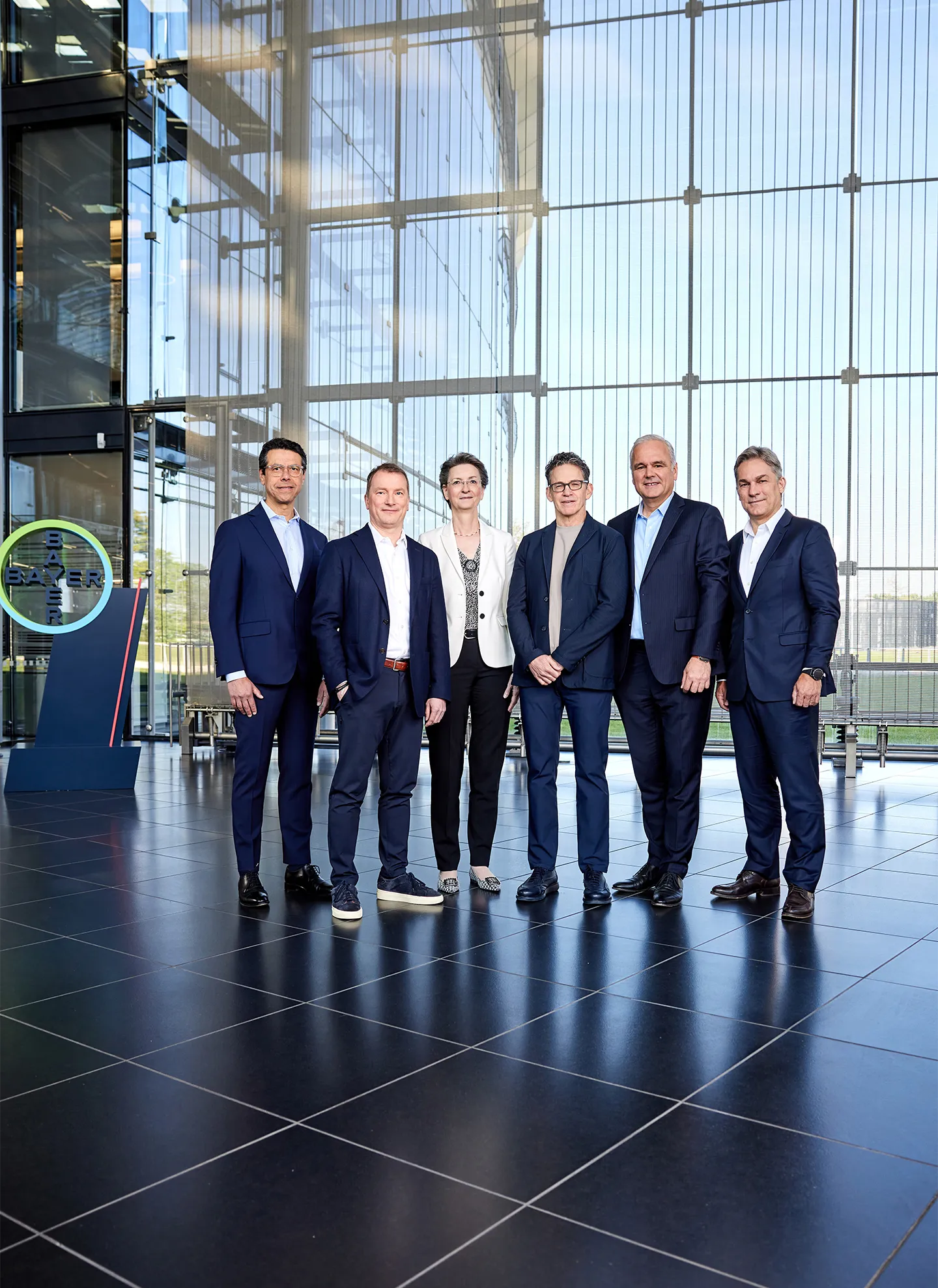 The board of Bayer, consisting of five men and one woman, is posing in business attire for a group photo inside a modern building in front of a glass wall, with a lawn visible behind it. The Bayer Group logo is visible on the left side.