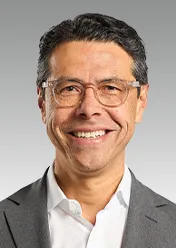Julio Triana is wearing a white shirt, a gray blazer, and glasses, smiling at the camera.