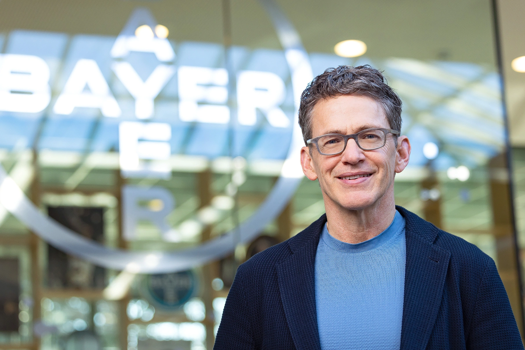 Bill Anderson is wearing a light blue T-shirt with a blue blazer and glasses, smiling at the camera. In the background, part of the Bayer logo and a glass-walled office can be seen.