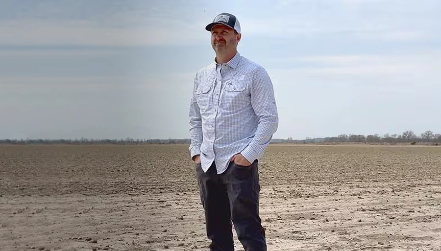 Ein Mann mit hellem, gestreiftem Hemd, dunkler Hose und einem schwarz-weißen Basecap schaut auf einem Feld in die Ferne.
