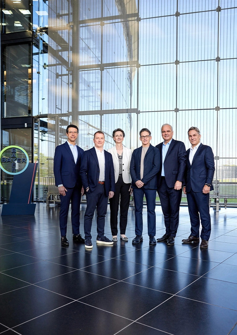 Der Vorstand von Bayer, bestehend aus fünf Männern und einer Frau, posiert in Businesskleidung für ein Gruppenfoto in einem modernen Gebäude vor einer Glaswand, hinter der eine Wiese zu sehen ist. Auf der linken Seite ist das Logo der Bayer-Gruppe sichtbar.