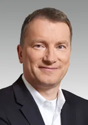 Wolfgang Nickl trägt ein weißes Hemd und ein schwarzes Sakko und lächelt in die Kamera.