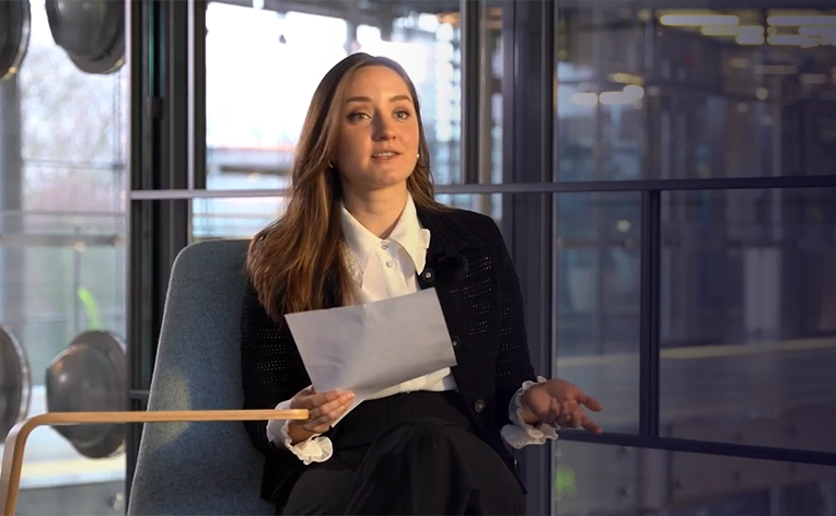 A woman with long brown hair sits in a chair in a modern office, holding a sheet of paper while speaking and gesturing with one hand. She is wearing a white collared shirt under a black cardigan, with large windows and a blurred office interior in the background.
